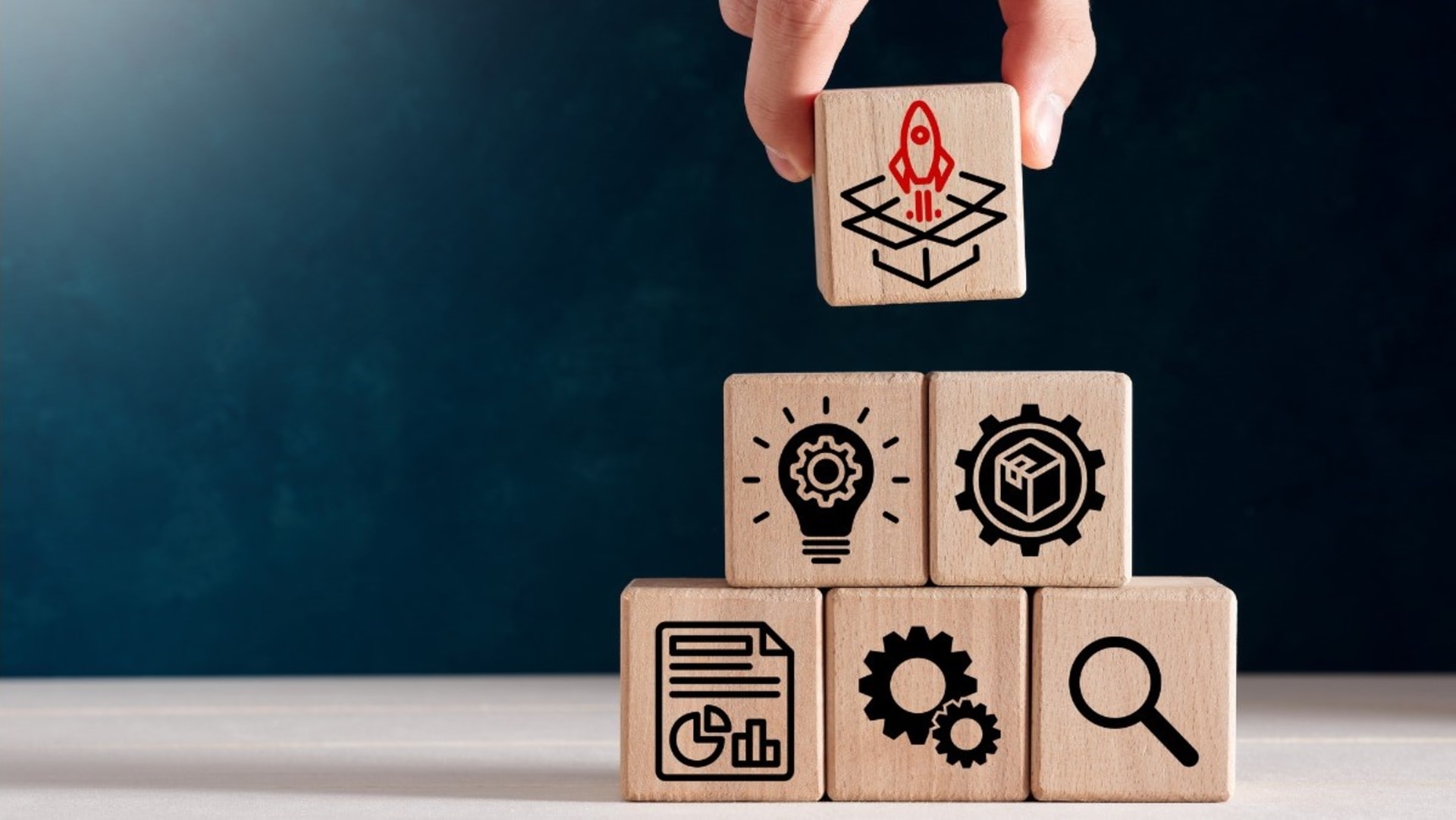 Hand placing a wooden block with a rocket illustration on top of other blocks showcasing icons of a light bulb, gear, magnifying glass, document, and 3D cube against a dark blue background.