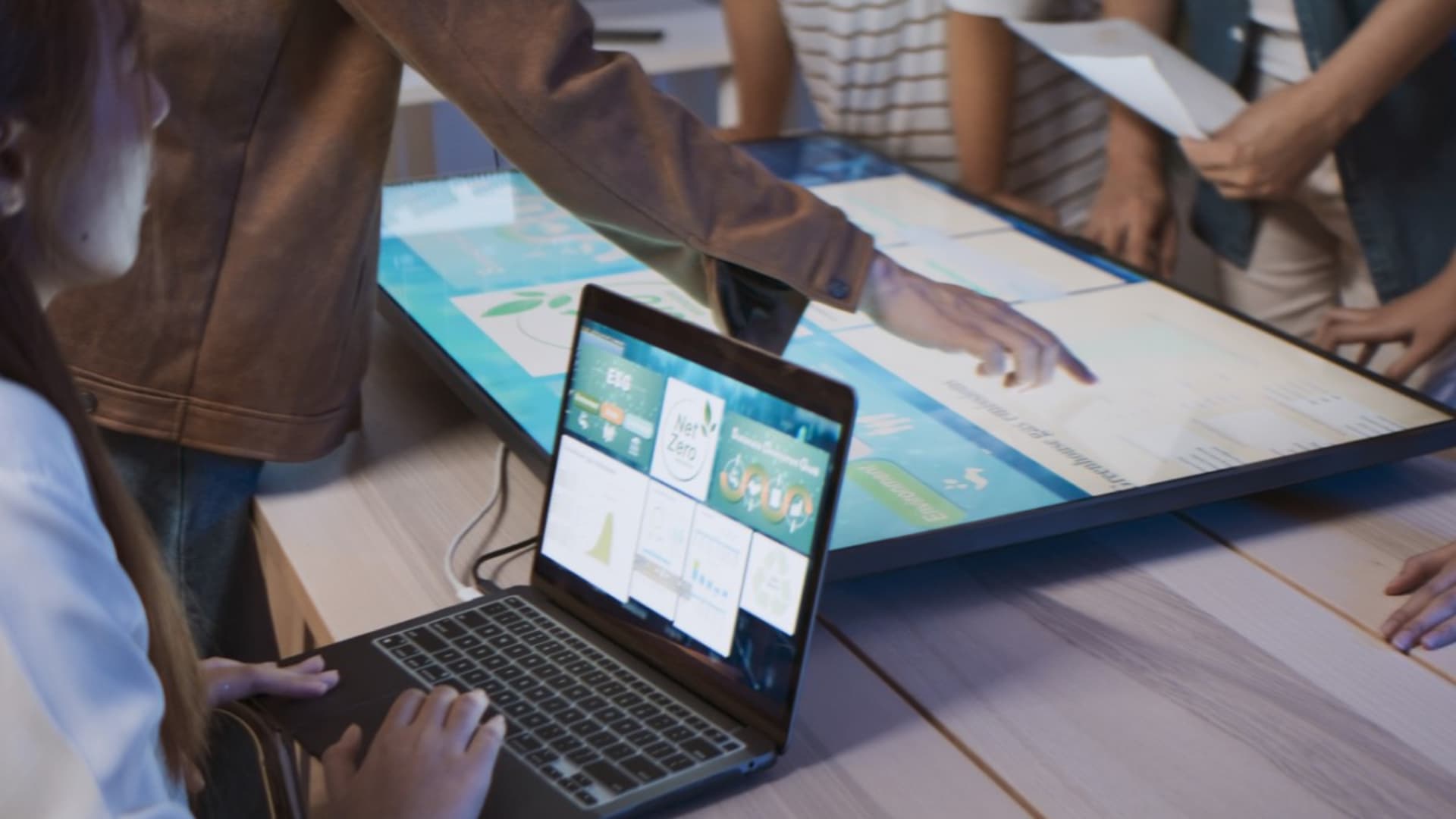 Team members interact with a large touch screen displaying sustainability dashboards while another person operates a laptop showing similar data visualizations.